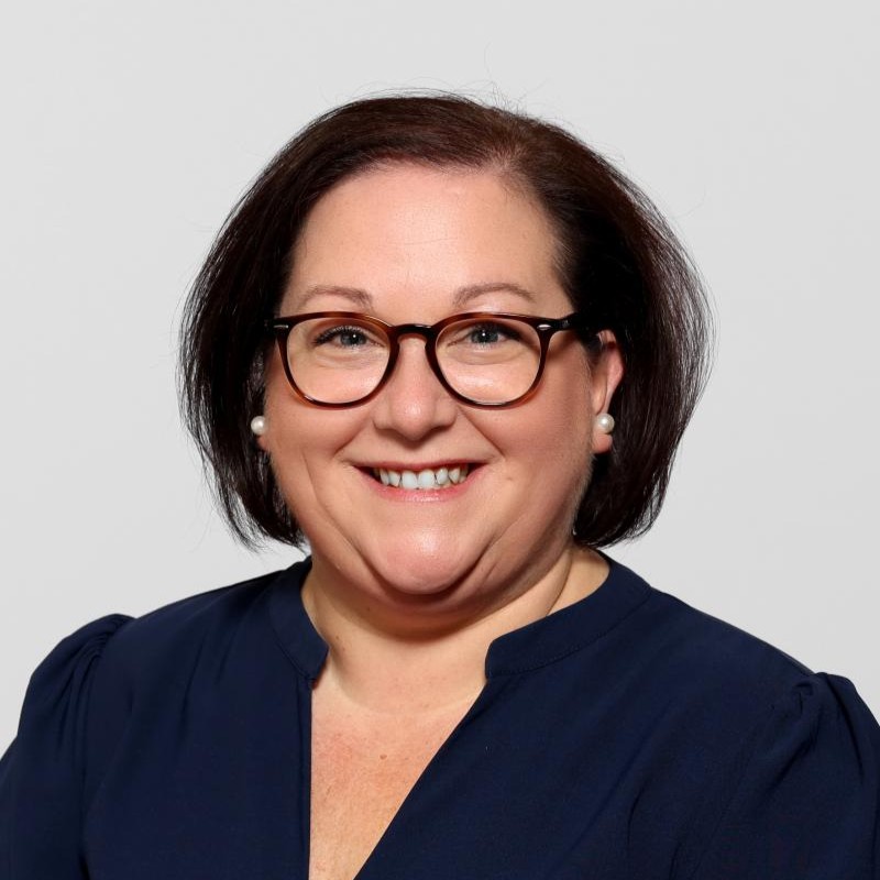 A professional headshot of a smiling woman with dark hair and glasses, wearing a dark top.